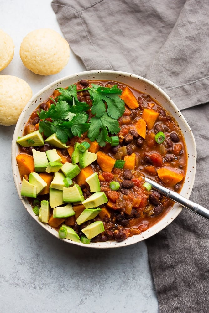 bowl of chili with sweet potatoes, avocado and cilantro on top, grey towel and background