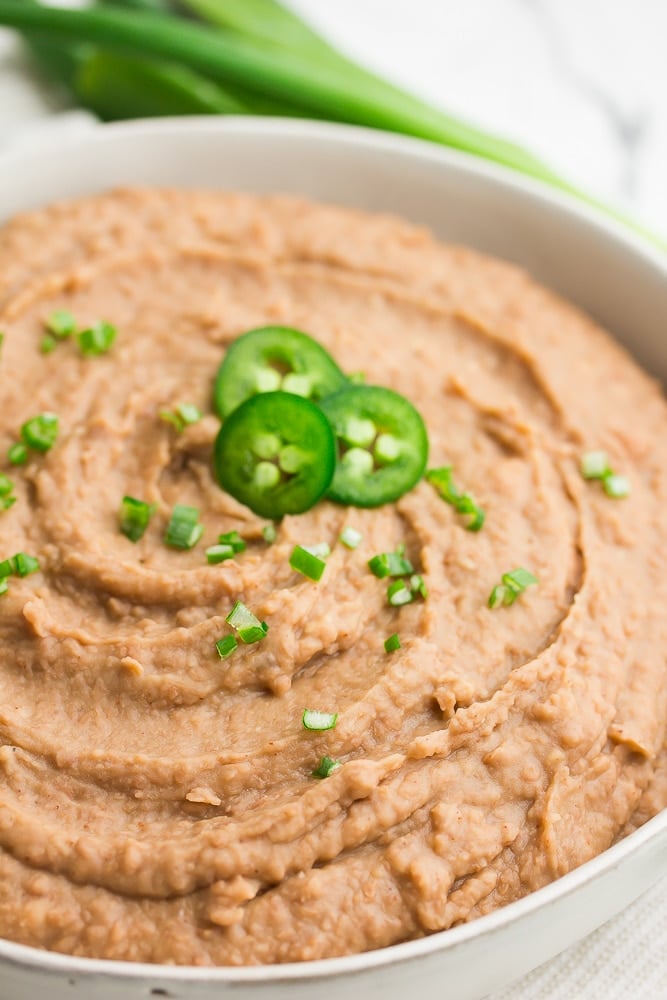 close up of a white bowl filled with refried beans with diced jalapenos and green onions on top.