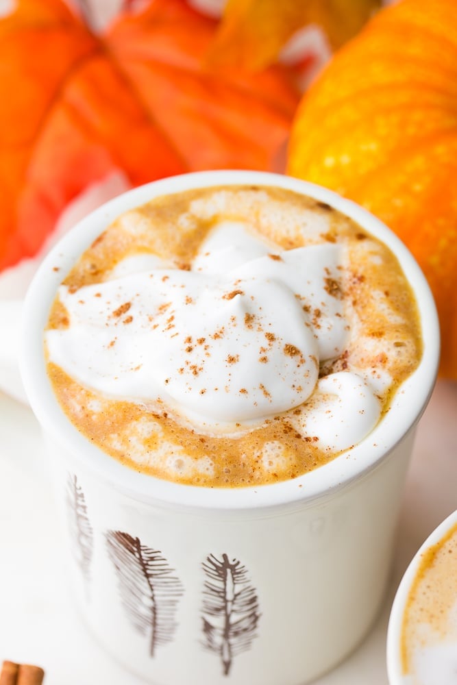 white cup with brown leave with vegan pumpkin spice latte, pumpkins in background