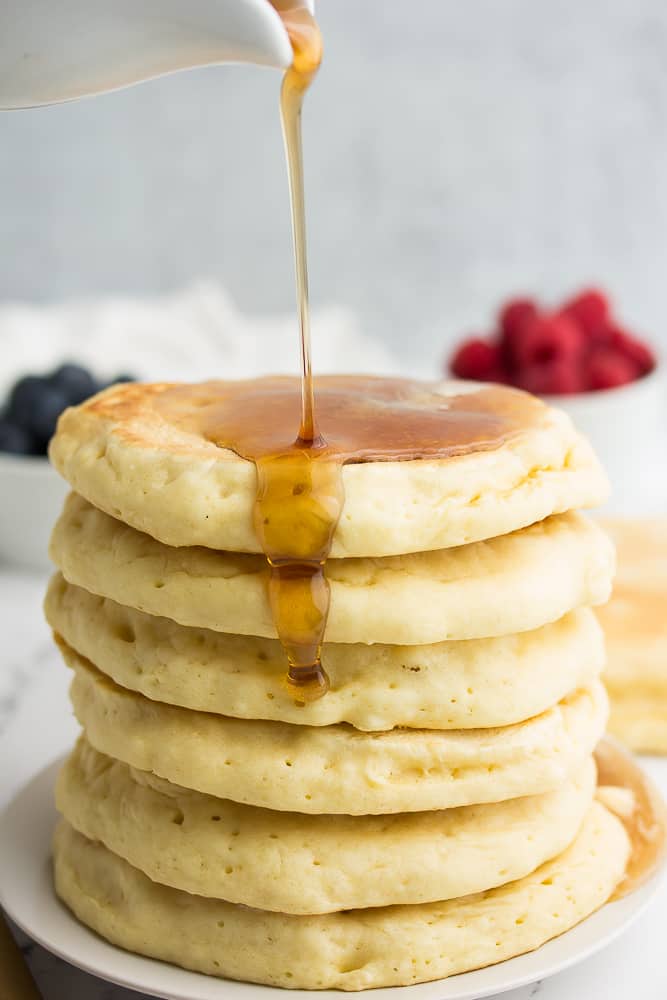 syrup being drizzled over stack of pancakes, berries and grey background