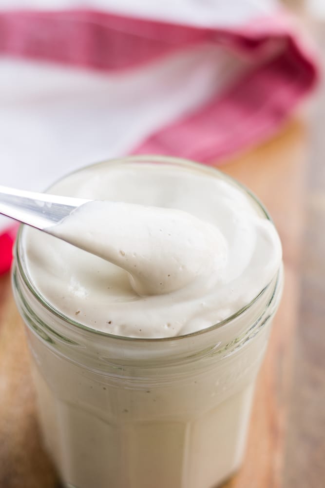 homemade vegan mayo in a glass jar with a small knife, red towel in background.