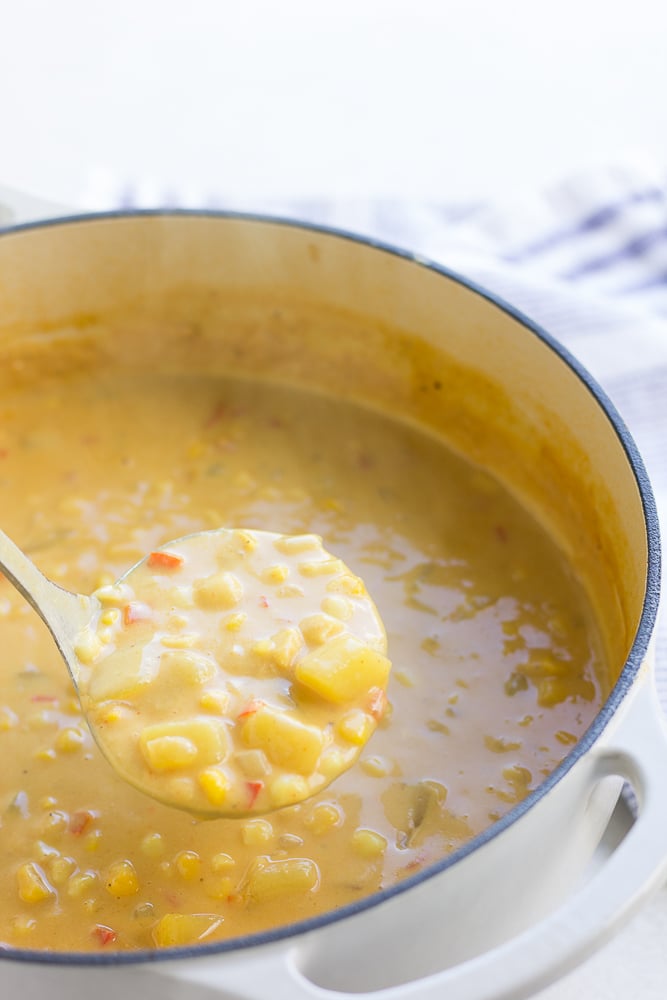 ladle held up with corn chowder out of a white pot.
