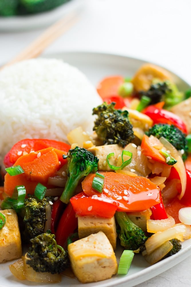 close up photo of tofu stir fry on a plate with rice and chopsticks.