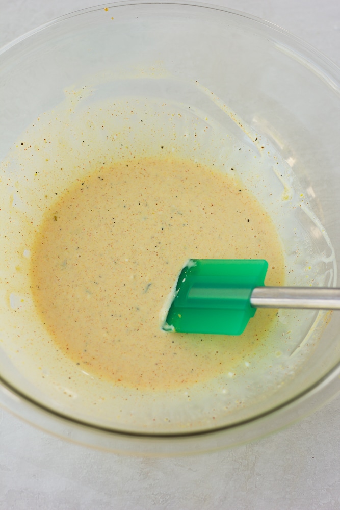 a green spatula stirring a yellow creamy sauce in a glass bowl.