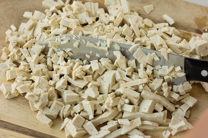 using a knife to chop tofu into small pieces on a wood cutting board.