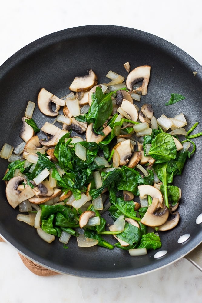 spinach and mushrooms and onions in a black pan being cooked