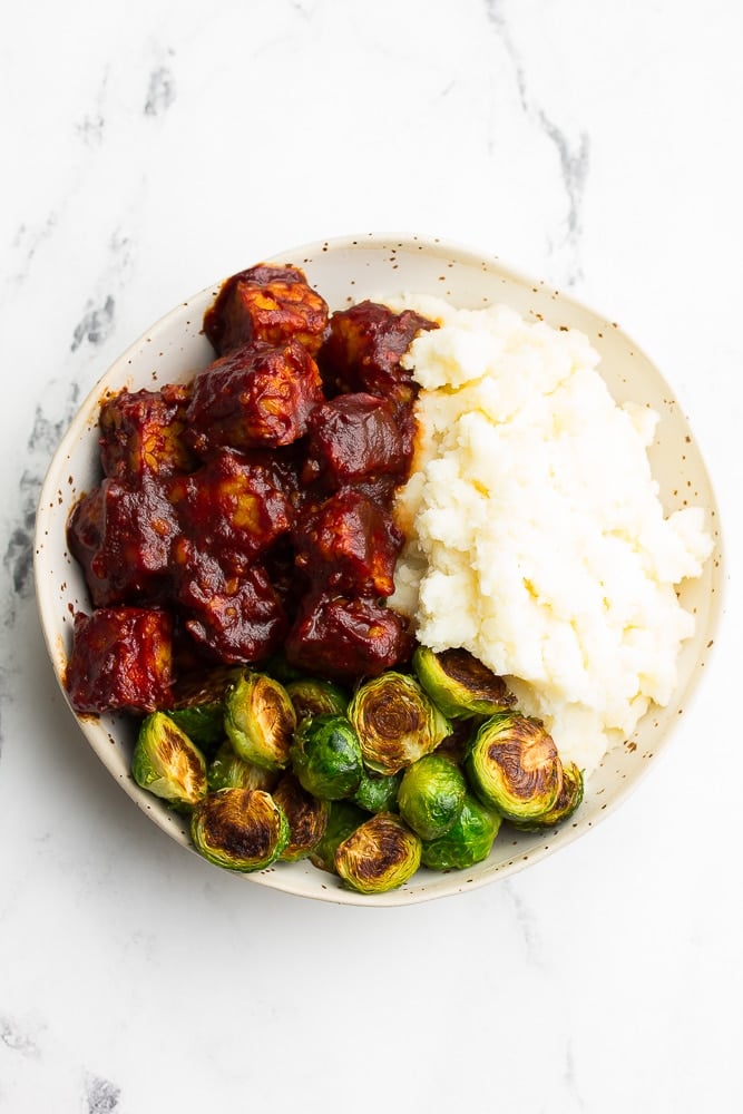 white bowl with bbq tempeh, mashed potatoes and brussels, white background