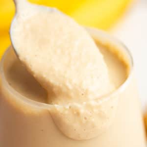 square image of a brown smoothie with bananas in background