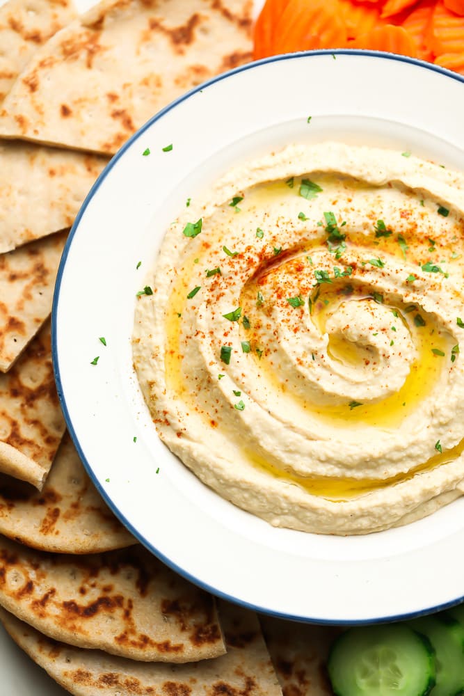 white bowl of hummus with pita bread and veggies around it