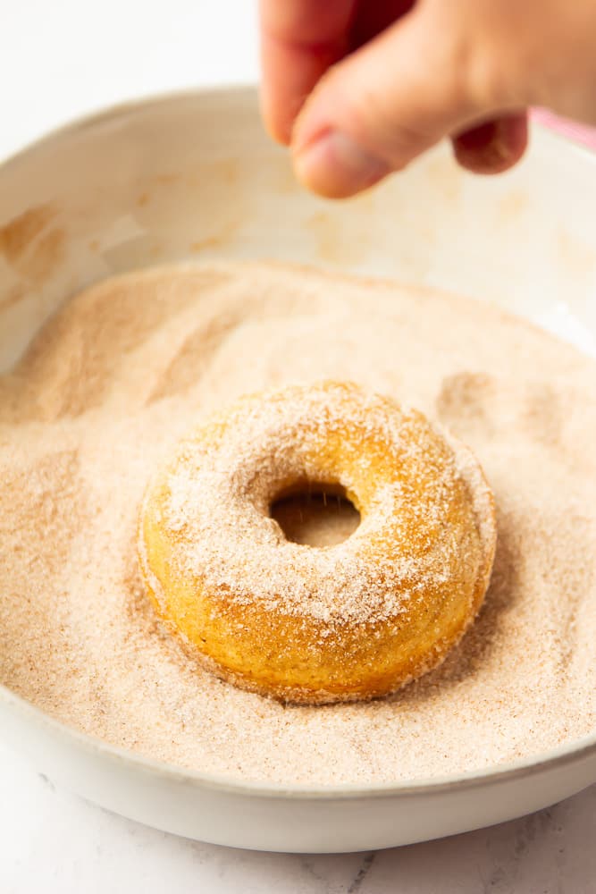 a donut being dipped and sprinkled in cinnamon sugar