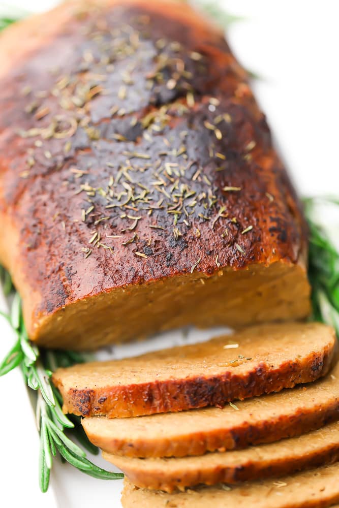 a vegan turkey loaf, browned on the top with herbs, partially sliced on a white plate