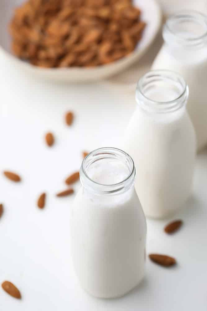 two glass milk bottles holding white almond milk, almonds in background