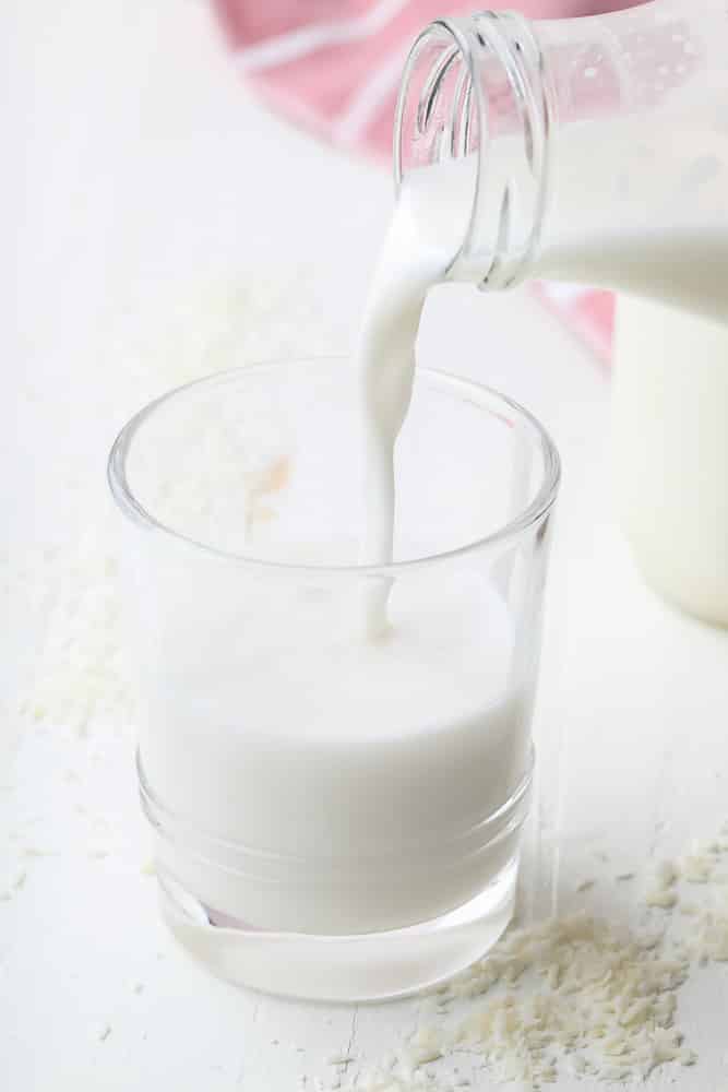 cup of milk being poured from a milk jug with coconut around it