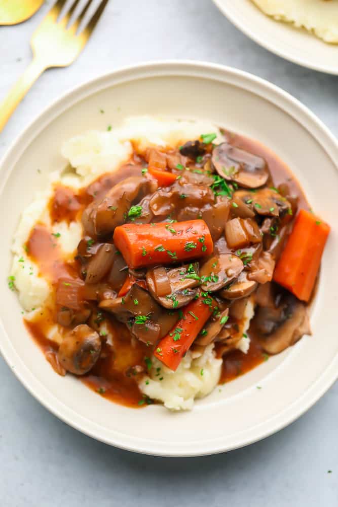 Close up of a white bowl with mashed potatoes and a mushroom and vegetable stew on top