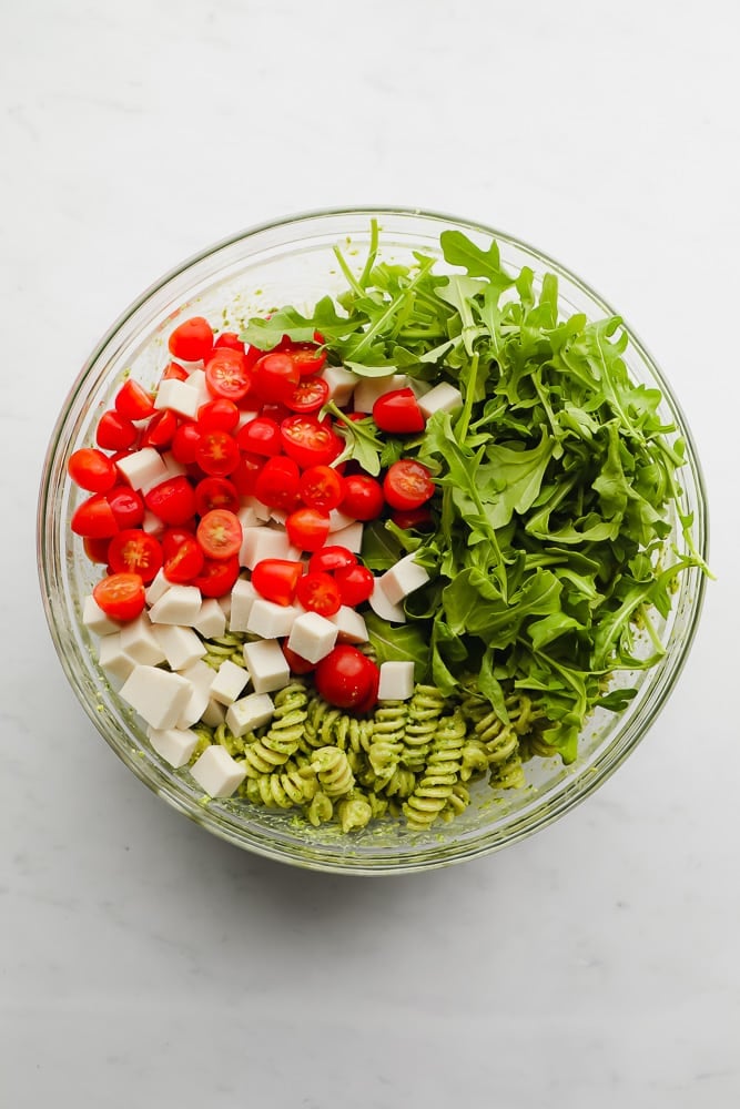 glass bowl filled with pesto pasta, arugula, cherry tomatoes, and cubed vegan cheese