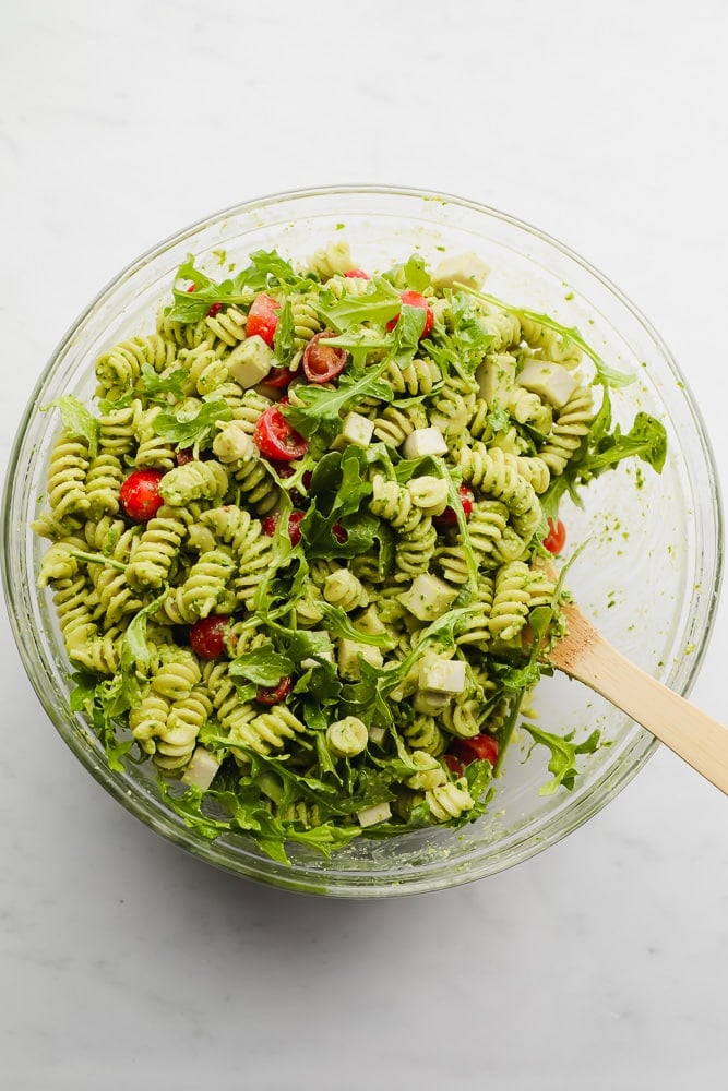 wooden spoon mixing a green pasta salad in a glass bowl