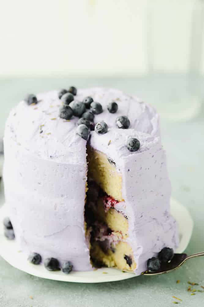 tall lavender cake on white plate with a slice being removed