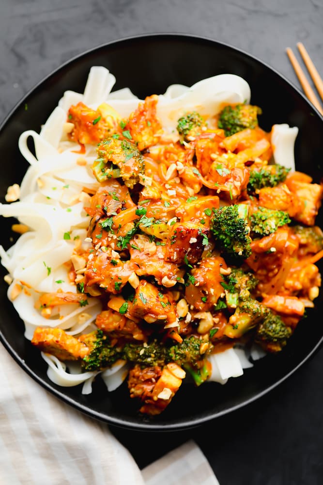 orange tempeh stir fry on top of white noodles in a black bowl