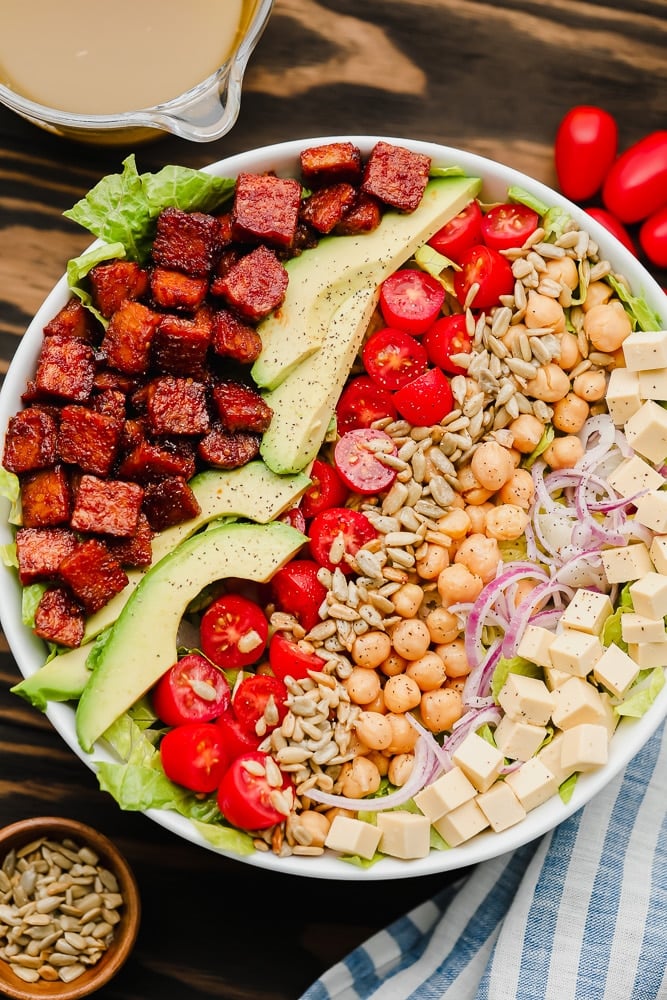 lines of vegan bacon, avocado, tomatoes, seeds, and beans on top of lettuce in a white bowl