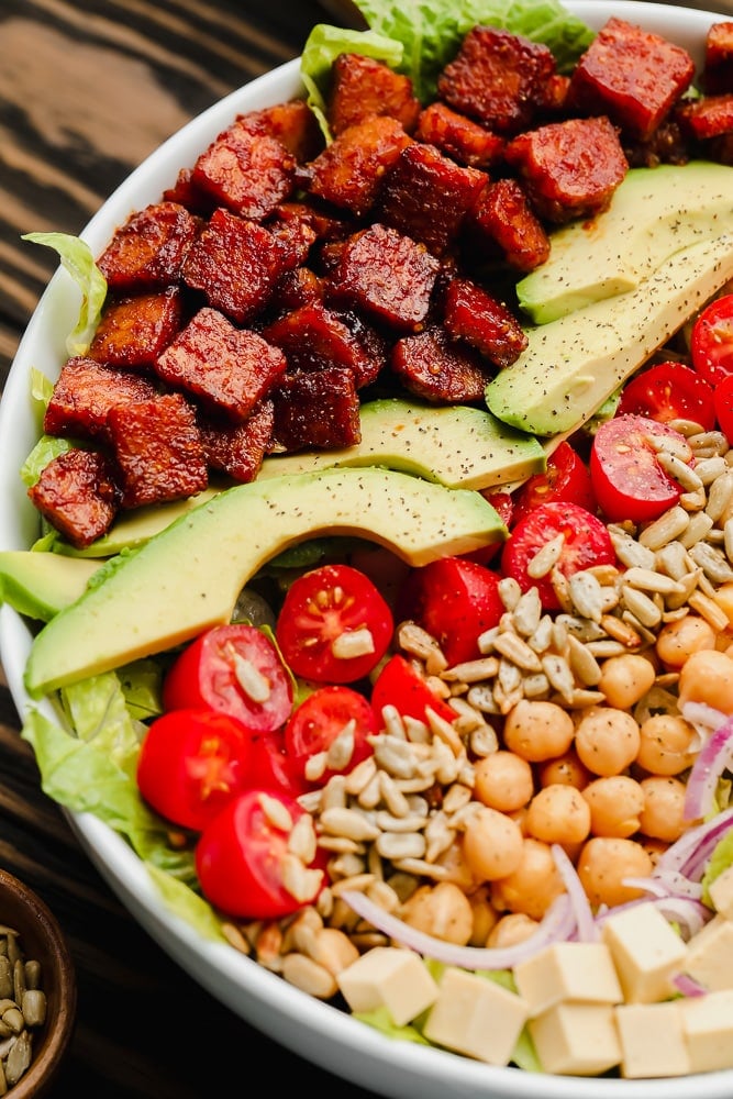 close up of lines of vegan bacon, avocado, tomatoes, seeds, and beans on top of lettuce in a white bowl