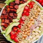 close up of lines of vegan bacon, avocado, tomatoes, seeds, and beans on top of lettuce in a white bowl