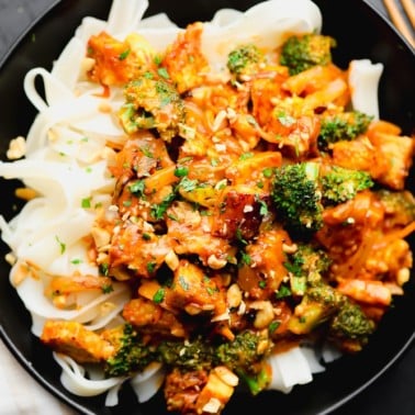 close up on a tempeh and broccoli stir fry on top of white noodles in a black bowl