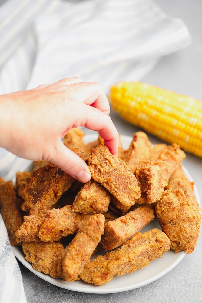 womans hand taking a piece of fried vegan chicken from a pile on a white plate