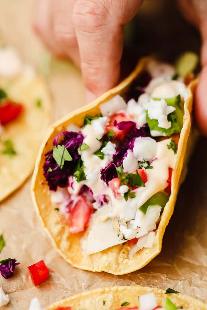 a hand picking up a corn tortilla filled with white sauce, tomatoes, and purple cabbage