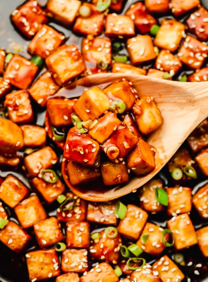 wooden spoon taking a scoop of sauce-covered tofu cubes out of a skillet