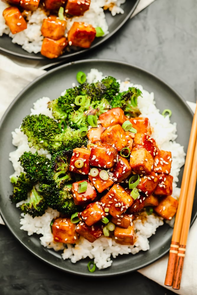 saucy brown tofu over white rice and cooked broccoli on a grey plate with chopsticks
