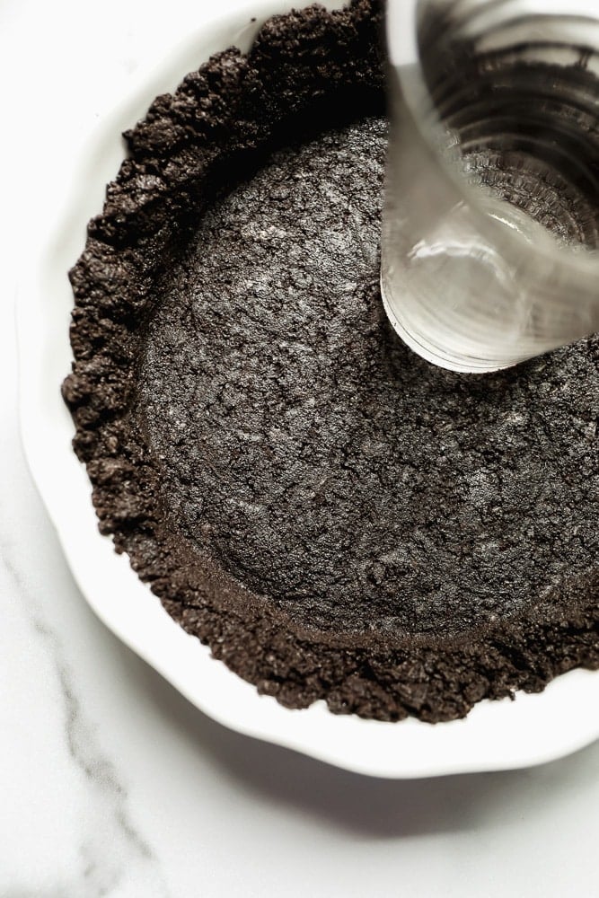 using a water glass to press a dark brown cookie crust into a white pie pan