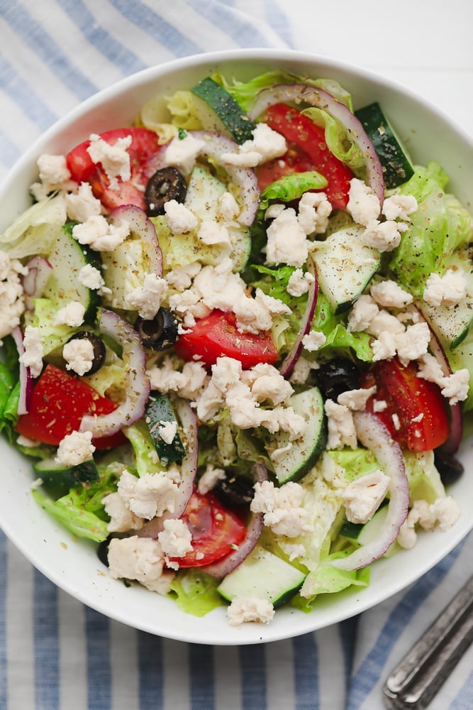 a Greek salad in a white bowl topped with crumbled white vegan feta cheese.