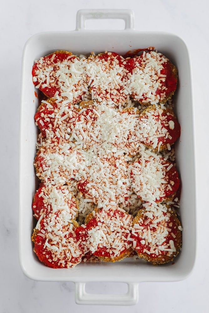 eggplant slices covered in red tomato sauce and white cheese in a white baking dish.