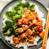 brown saucy tofu on a grey plate with white rice and broccoli