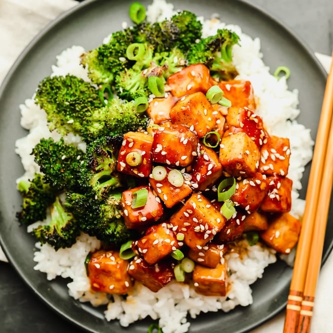 brown saucy tofu on a grey plate with white rice and broccoli