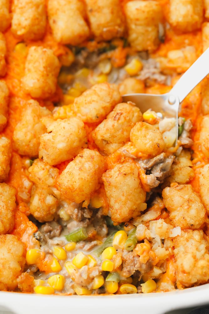 metal spoon scooping some baked tater tot casserole out of a white casserole dish.