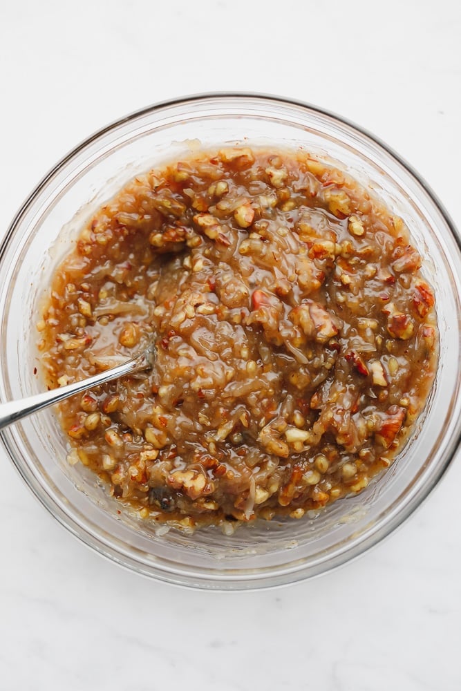 A spoon stirring a thick caramel sauce with pecans in a glass bowl.