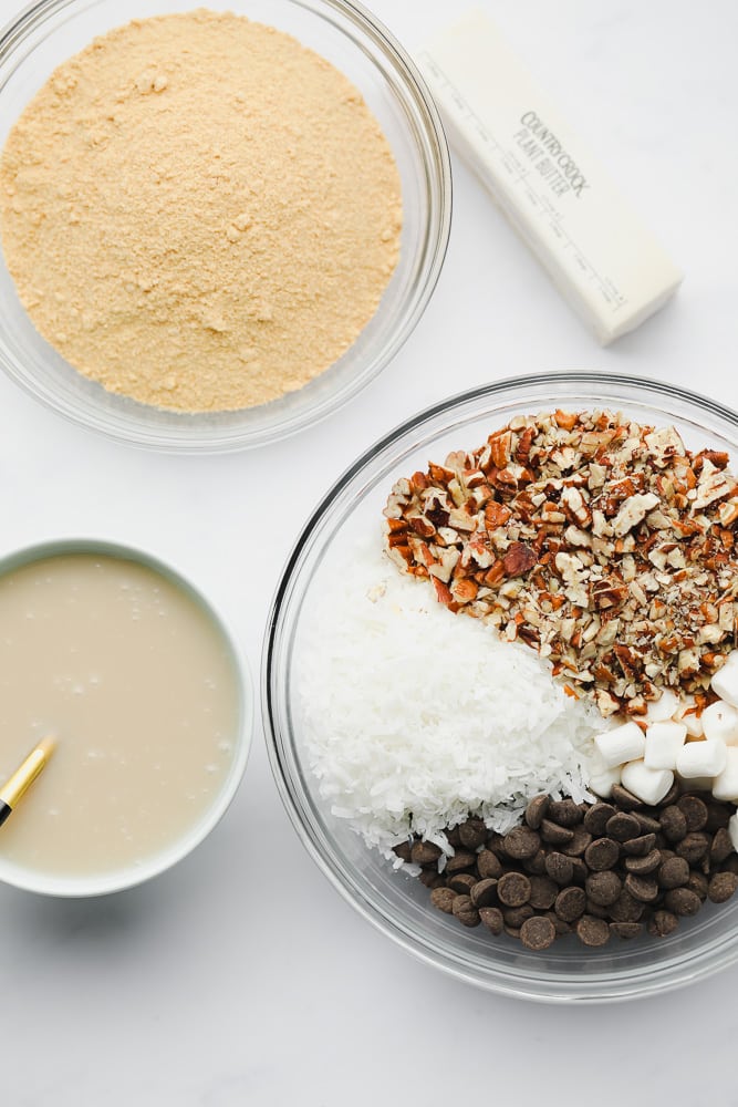 3 bowls filled with crushed graham crackers, nuts, chocolate, and coconut, and condensed milk.