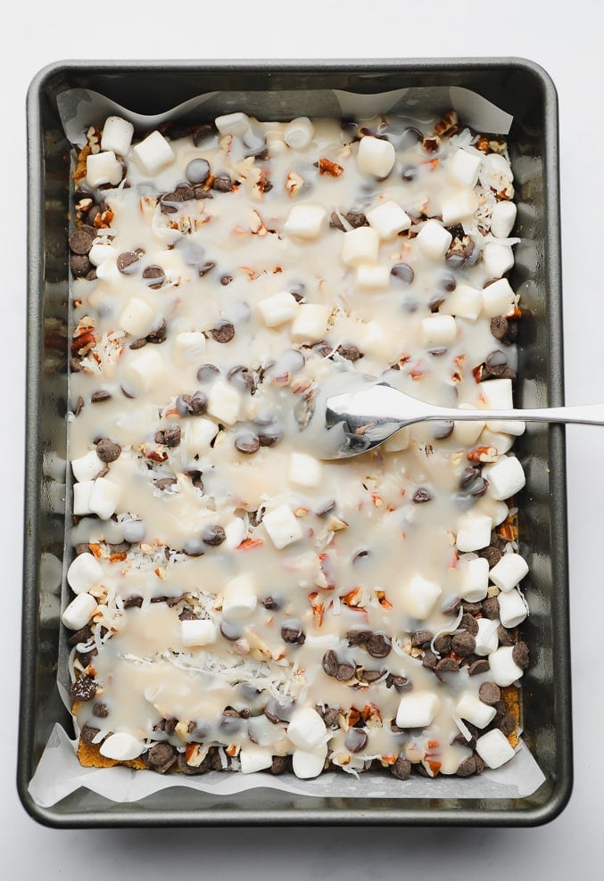 a spoon spreading condensed milk over layers of candy and chocolate in a metal baking pan.