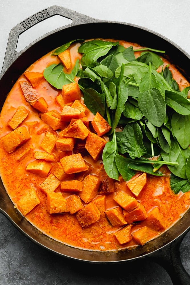 roasted sweet potato cubes and raw spinach on top of an orange liquid in a black skillet.
