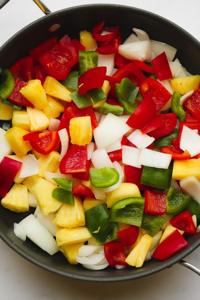 chopped onions, pineapple chunks, and red and green bell peppers in a large black skillet.