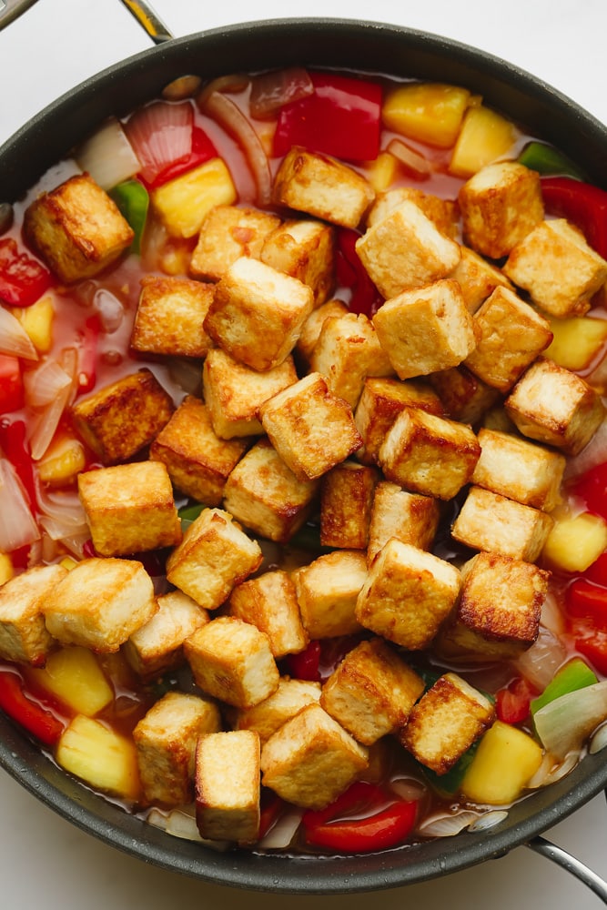 fried tofu cubes on top of chopped peppers, onions, and pineapple in a large black skillet.