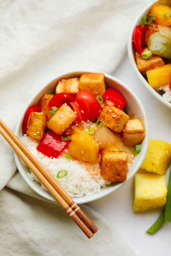 cooked sweet and sour tofu, pineapple, and peppers in a white bowl with rice and chop sticks.