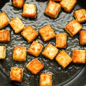 close up on golden brown tofu nuggets frying in oil in a black skillet.