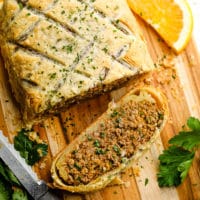 a sliced vegan beef wellington on a wood board surrounded by a knife, green herbs, and orange slices.