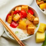 cooked sweet and sour tofu, pineapple, and peppers in a white bowl with rice and chop sticks.