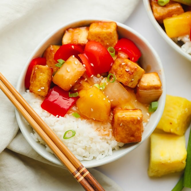 cooked sweet and sour tofu, pineapple, and peppers in a white bowl with rice and chop sticks.