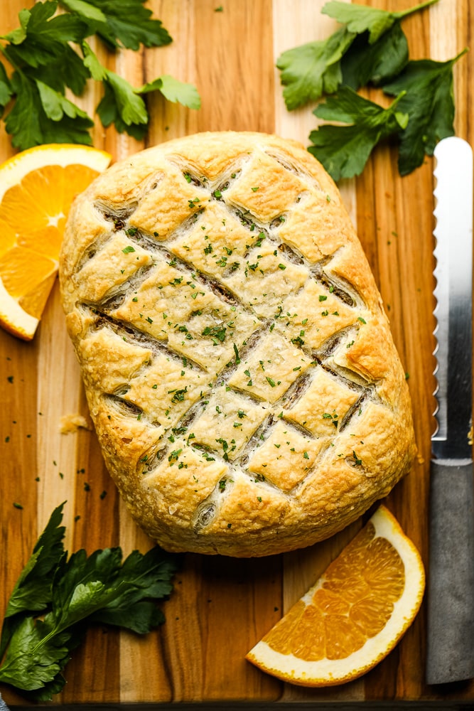 a baked vegan wellington on a wood board, surrounded by green herbs and orange slices.