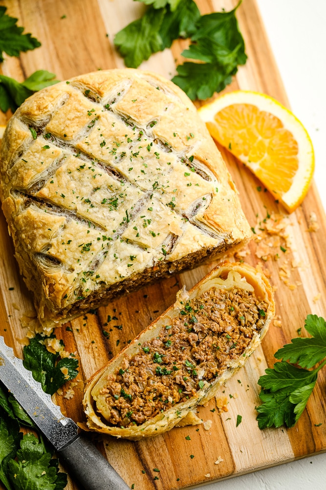 a sliced vegan beef wellington on a wood board surrounded by a knife, green herbs, and orange slices.