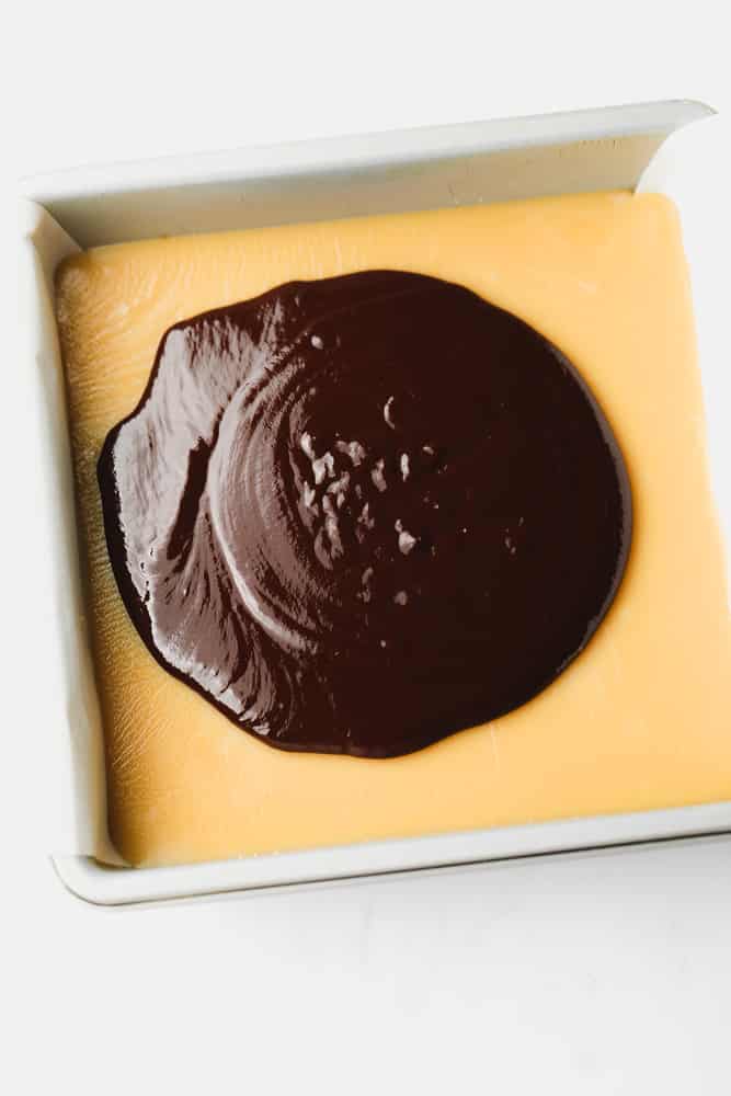 pouring melted chocolate over caramel in a metal baking dish.